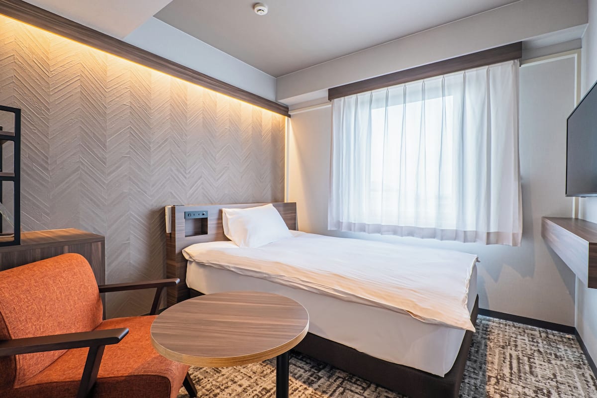 Ein Hotelzimmer mit Bett, Sessel, kleinem Tisch, Fernseher und einem Fenster mit Vorhängen.