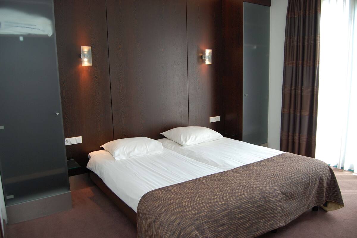 Une chambre d’hôtel moderne avec un grand lit, une tête de lit en bois foncé et deux lampes fixées au mur.
