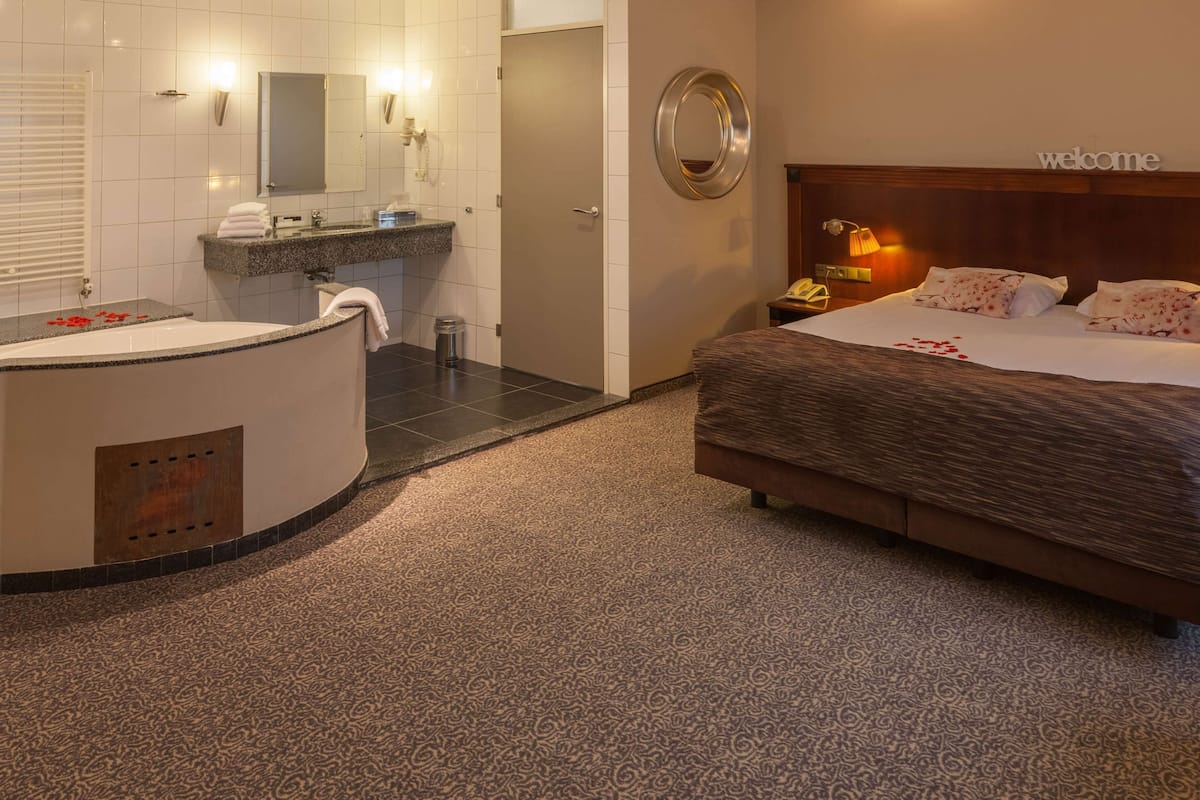 Une chambre d’hôtel avec un grand lit, une salle de bain avec une baignoire, un miroir rond et un panneau d’accueil.