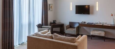 A hotel room with a sofa, two armchairs, a small table with a coffee maker, a TV mounted on the wall, and a desk with a 'welcome' sign.