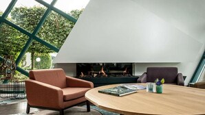 A modern interior with a fireplace, two armchairs, a round table, and a view of greenery through large windows. - Best Western Hotel Nobis Asten (Asten)