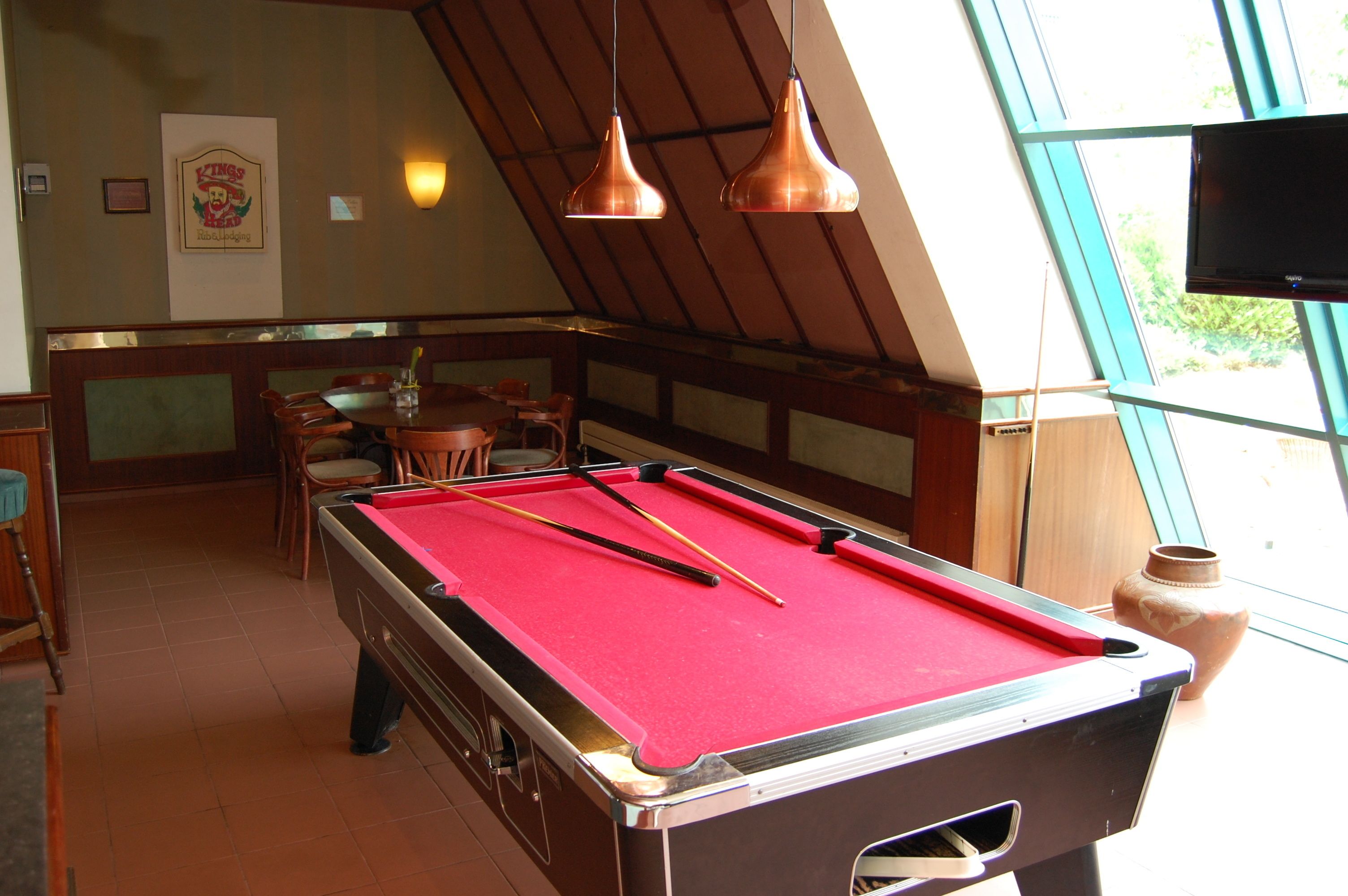 A pool table with red felt, a billiards cue, and a vase on a windowsill.