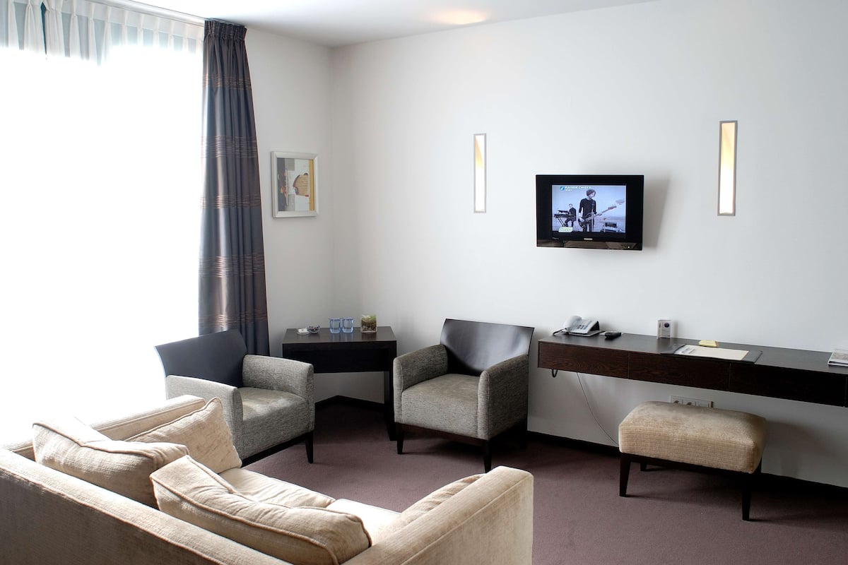 Une chambre d’hôtel moderne équipée d’un canapé, d’un fauteuil et d’un pouf, d’un bureau avec un téléviseur à écran plat et d’une grande fenêtre avec des rideaux.