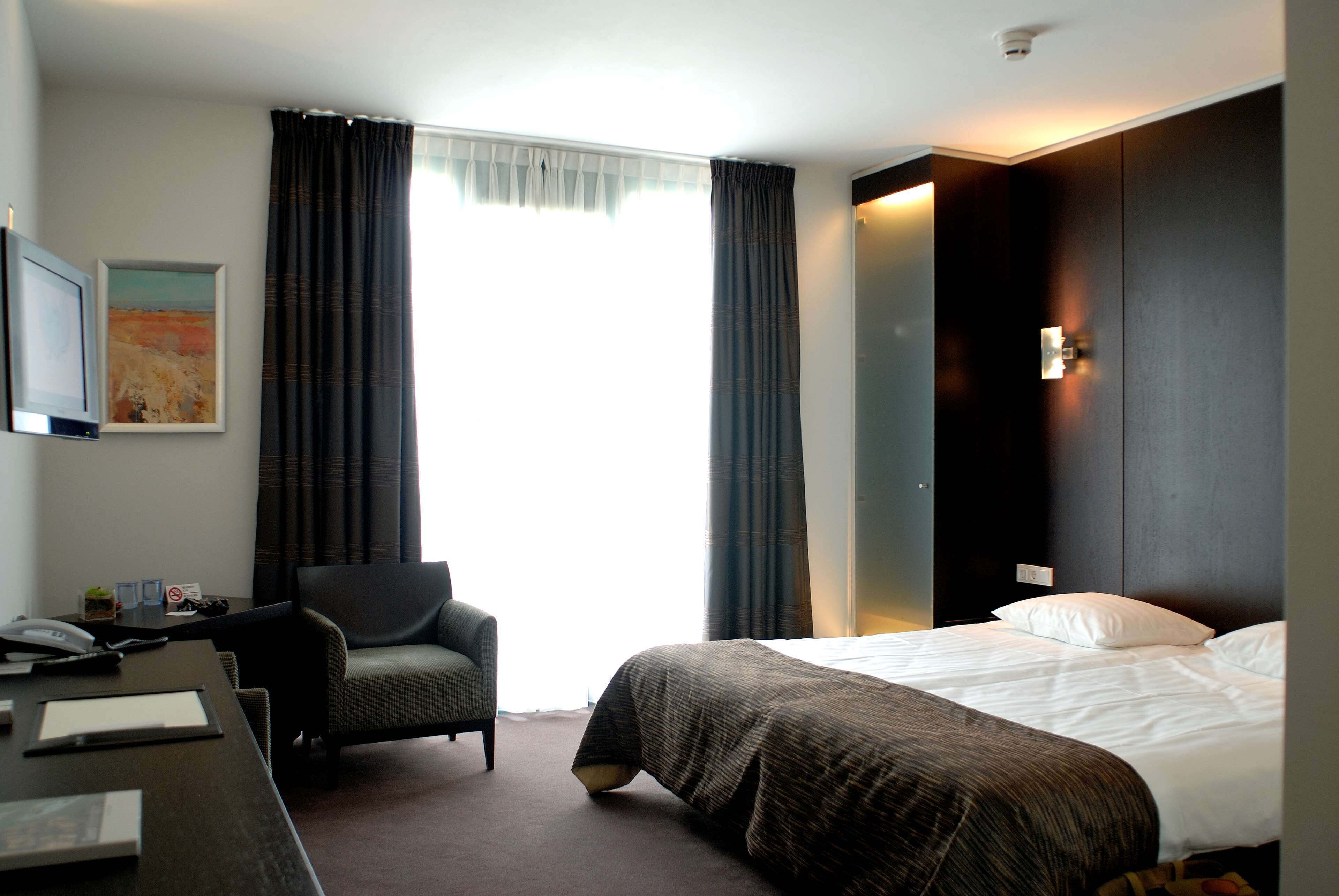 Una habitación de hotel con cama, escritorio, silla, un cuadro en la pared y una ventana con cortinas.