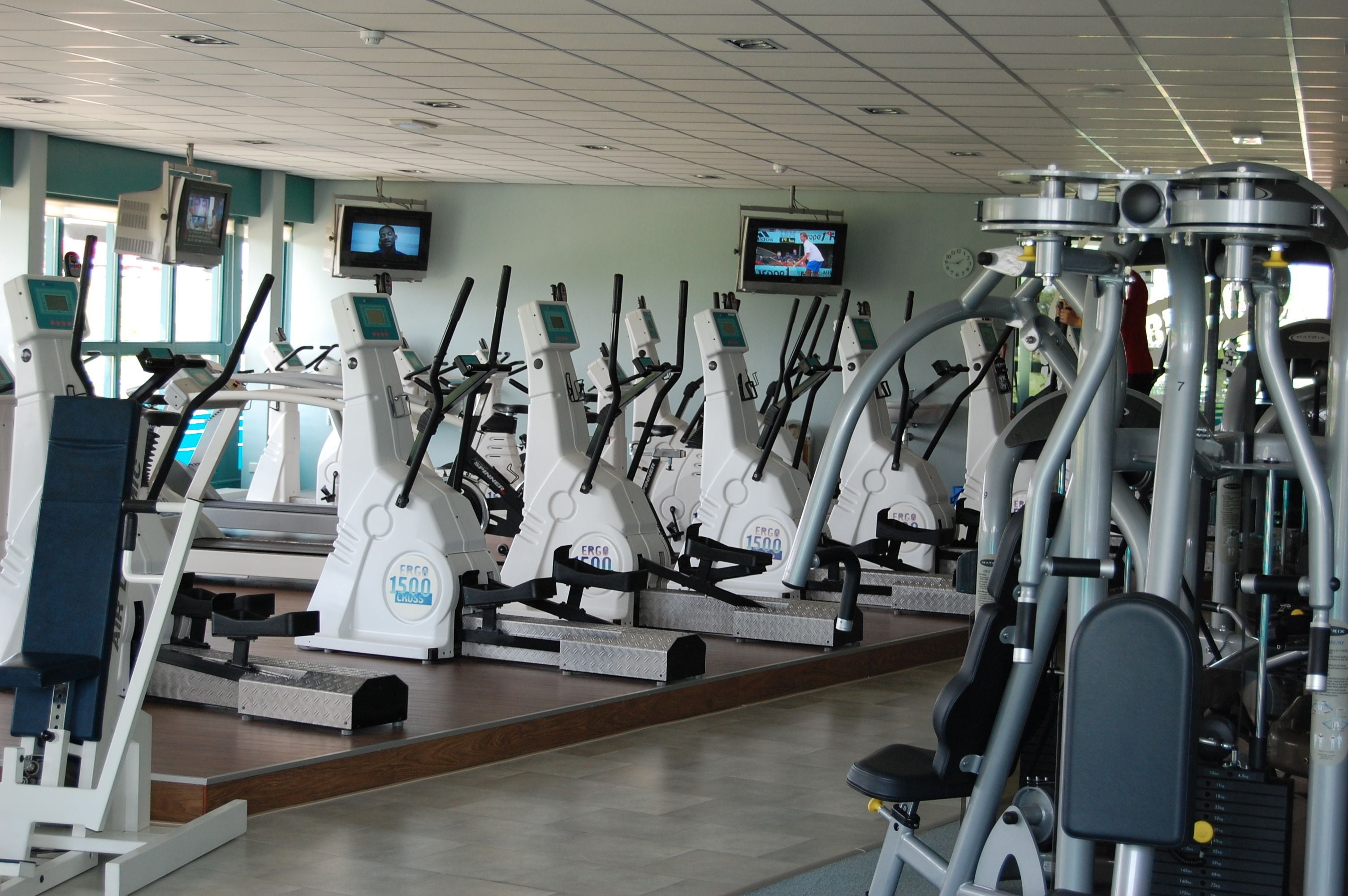 A gym with multiple cardio machines, including treadmills and elliptical trainers, arranged in rows.