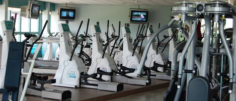 A gym with multiple cardio machines, including treadmills and elliptical trainers, arranged in rows.