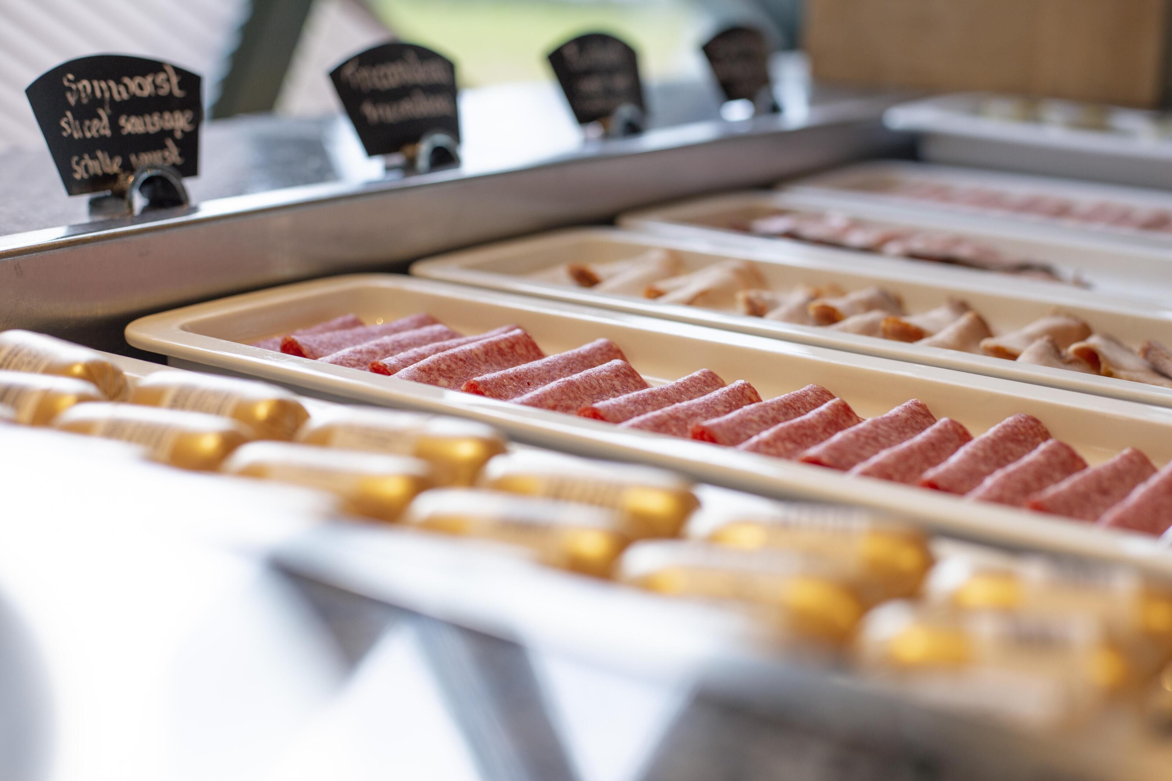 Exposição de alimentos com diversos tipos de carne fatiada e uma placa indicando 'Spitzwurst, salsicha fatiada'.