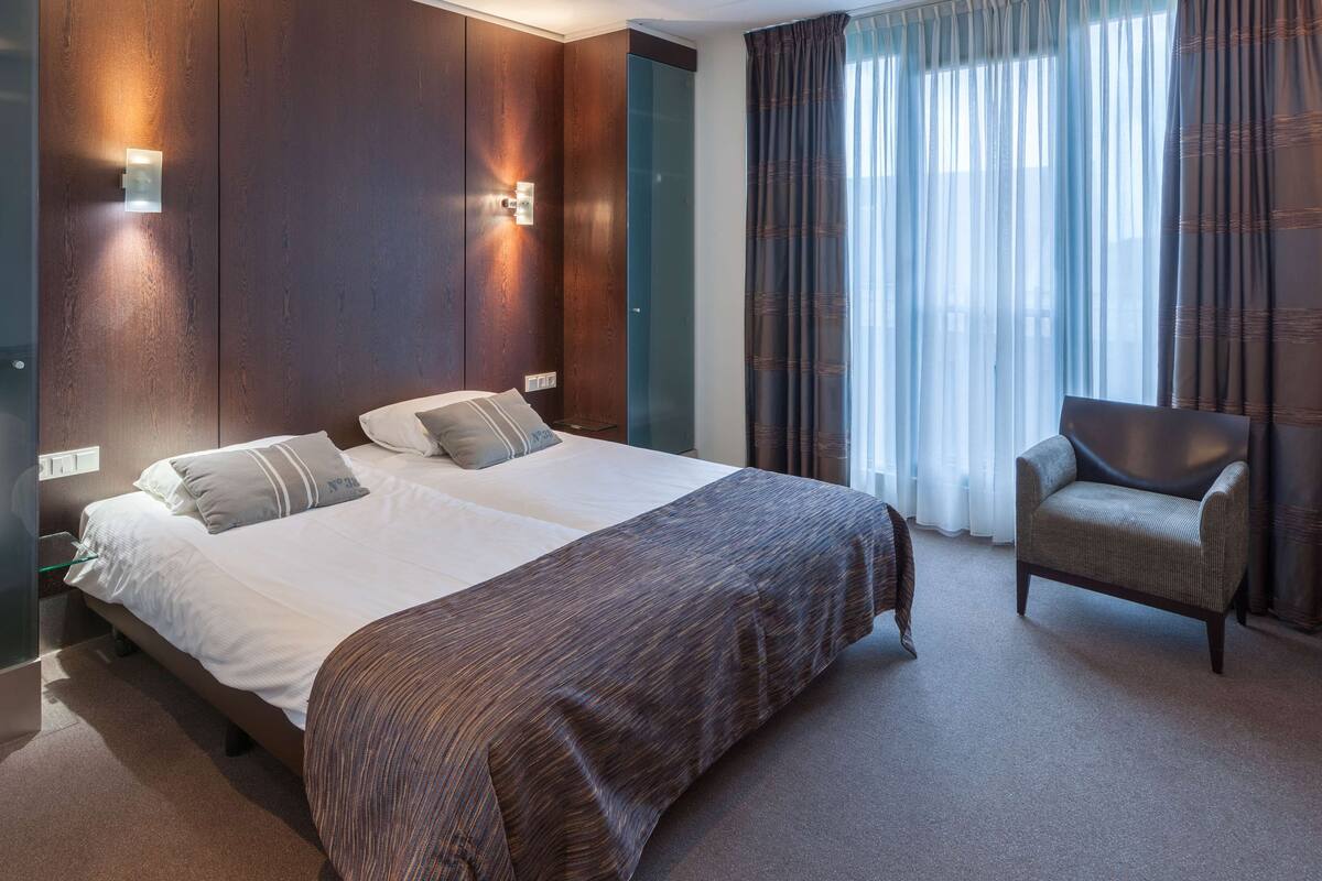 Une chambre d’hôtel avec un grand lit, une chaise et une fenêtre avec des rideaux.