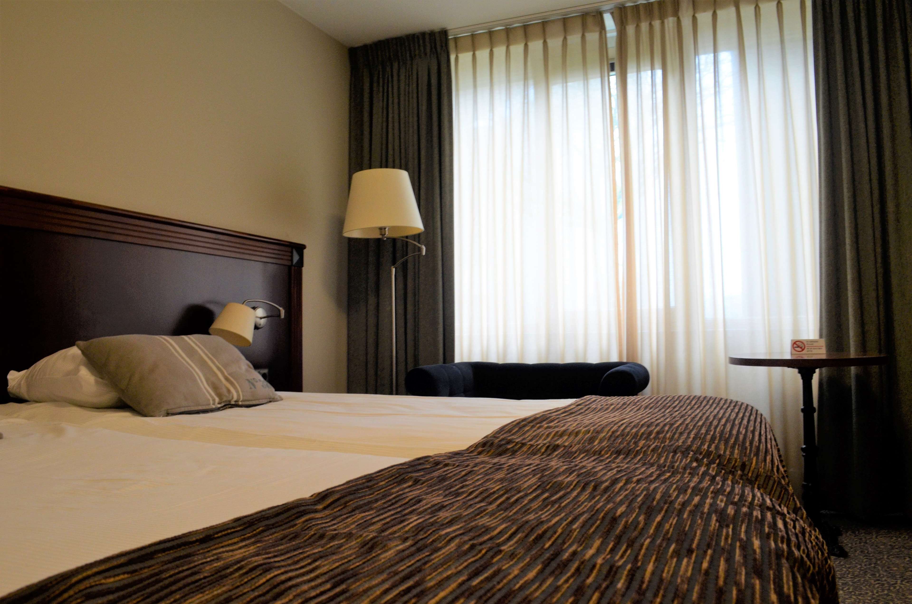 A hotel room with a large bed, a bedside lamp, a chair, and a small table.