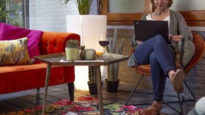 A woman sitting on a chair with a laptop, a red sofa, a small table with a plant, and a cactus in the background.