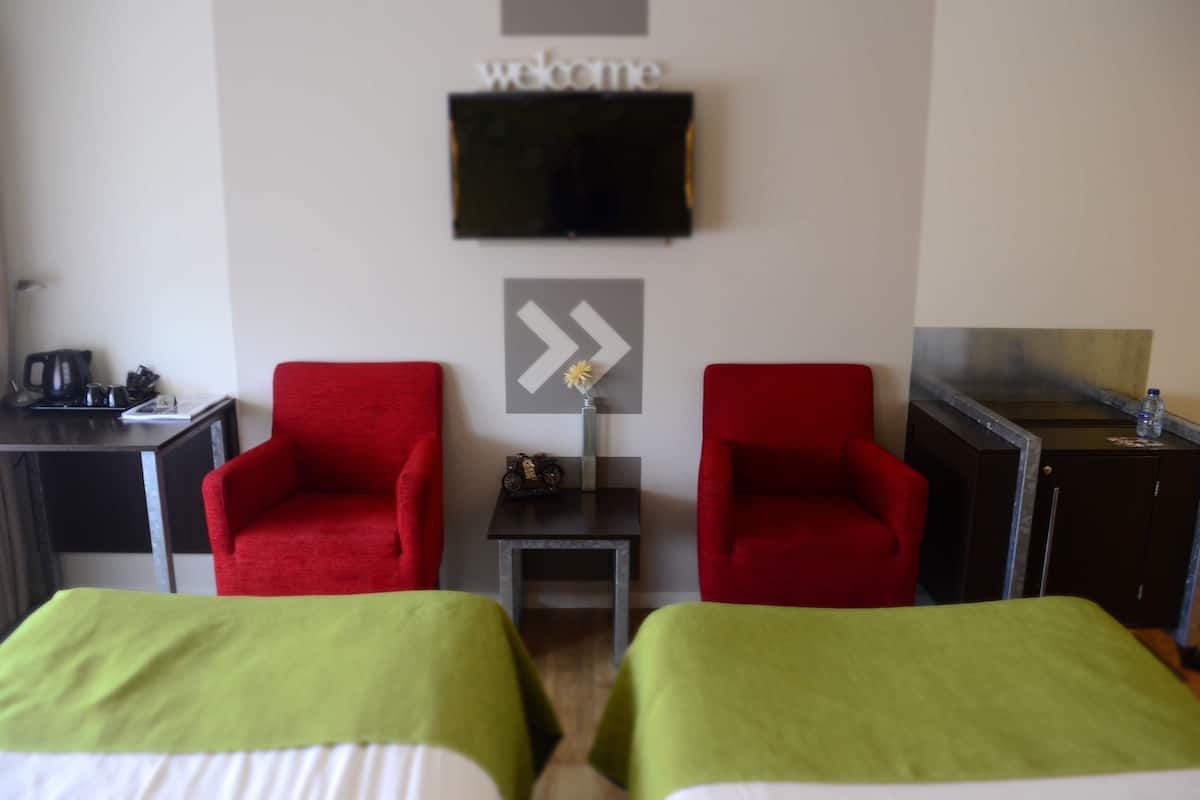 Habitación de hotel con dos camas individuales, un escritorio con cafetera, dos sillones rojos, una mesita con un florero, un televisor montado en la pared y un cartel que dice 'welcome'.