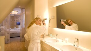 A hotel bathroom with a large mirror, two sinks, and a bed visible through a curtain.
