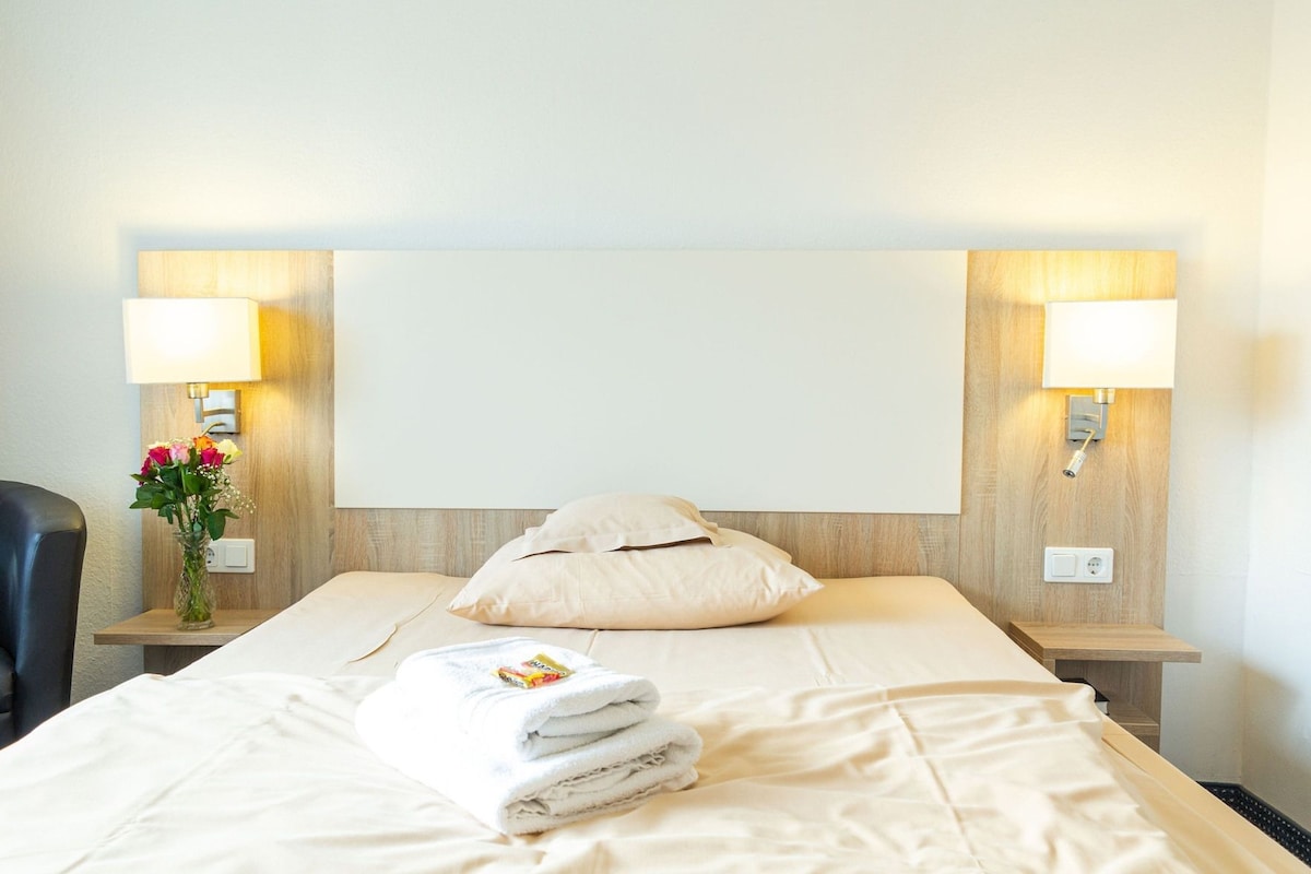 Een hotelkamer met een bed, twee bedlampen, een nachtkastje met een bloevas en een bedieningspaneel aan de muur.