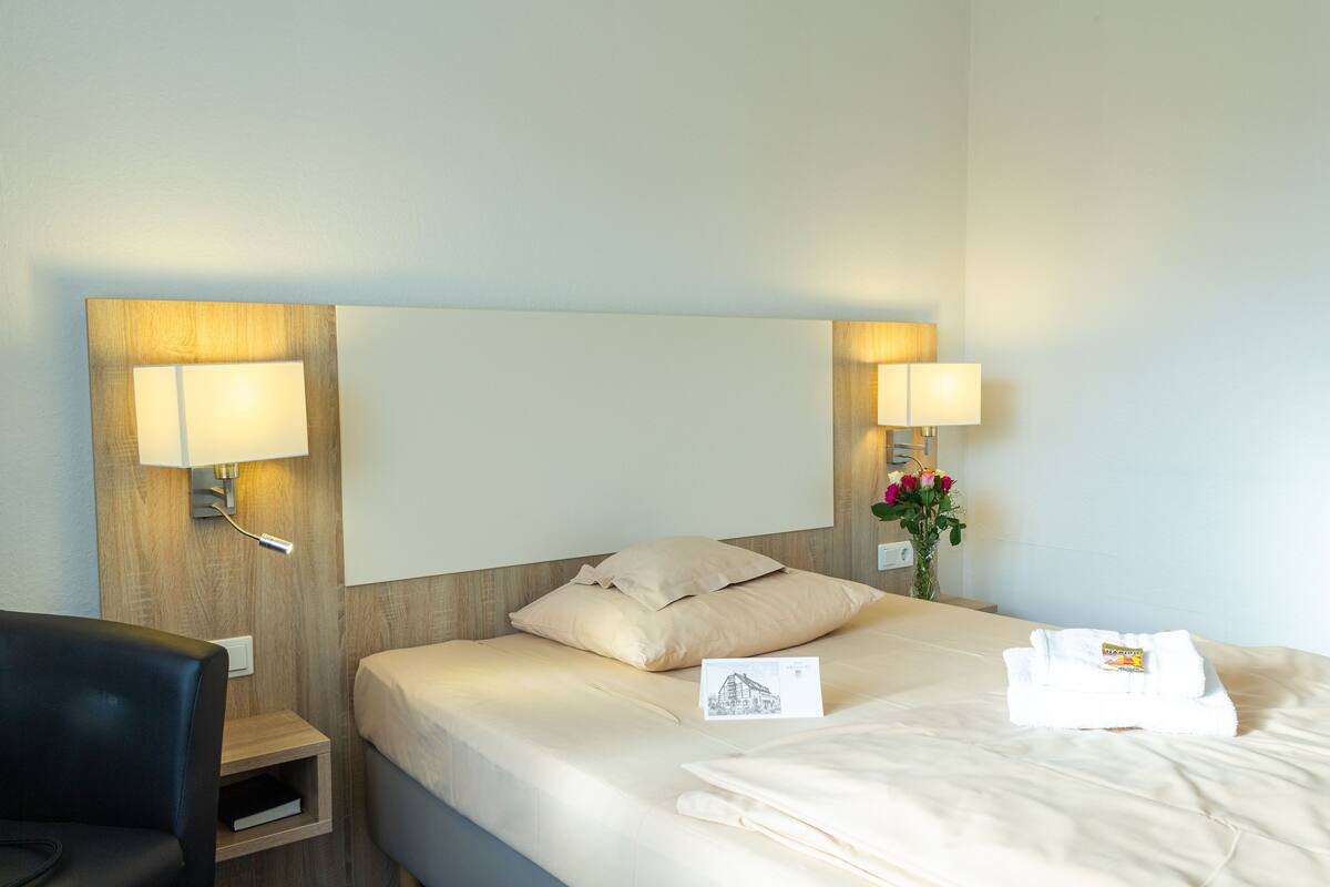 Een hotelkamer met een bed, twee bedlampen, een nachtkastje, een stoel en een vaas met bloemen.