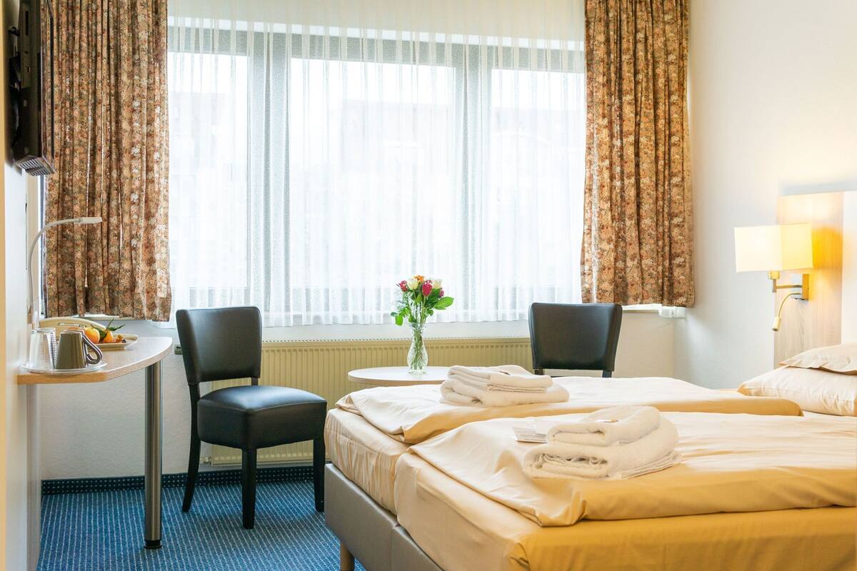 Een hotelkamer met twee bedden, een bureau, stoelen, een vaas met bloemen en een raam met gordijnen.