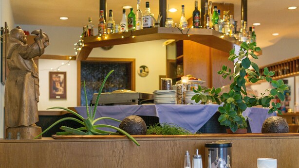 A restaurant interior with a wooden bench, a statue, a potted plant, and a bar area with bottles. - Hotel Kelkheimer Hof (Kelkheim)