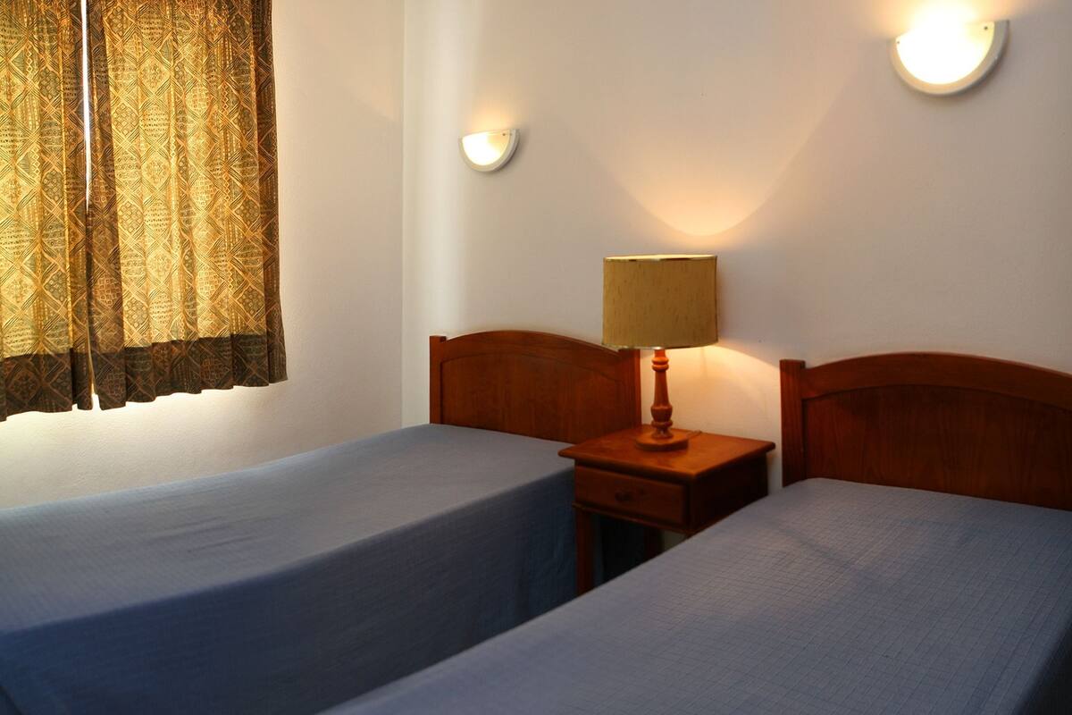 A room with two single beds, a nightstand with a lamp, and a wall-mounted light fixture.