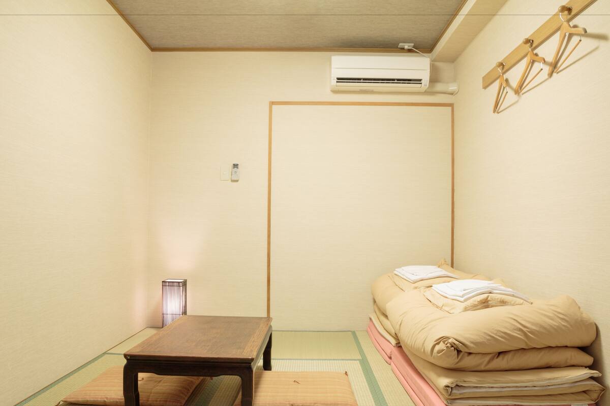 Ein Zimmer mit Tatami-Boden, einem niedrigen Holztisch, einem Bett mit Kissen, einer Klimaanlage und einem hölzernen Kleiderständer.