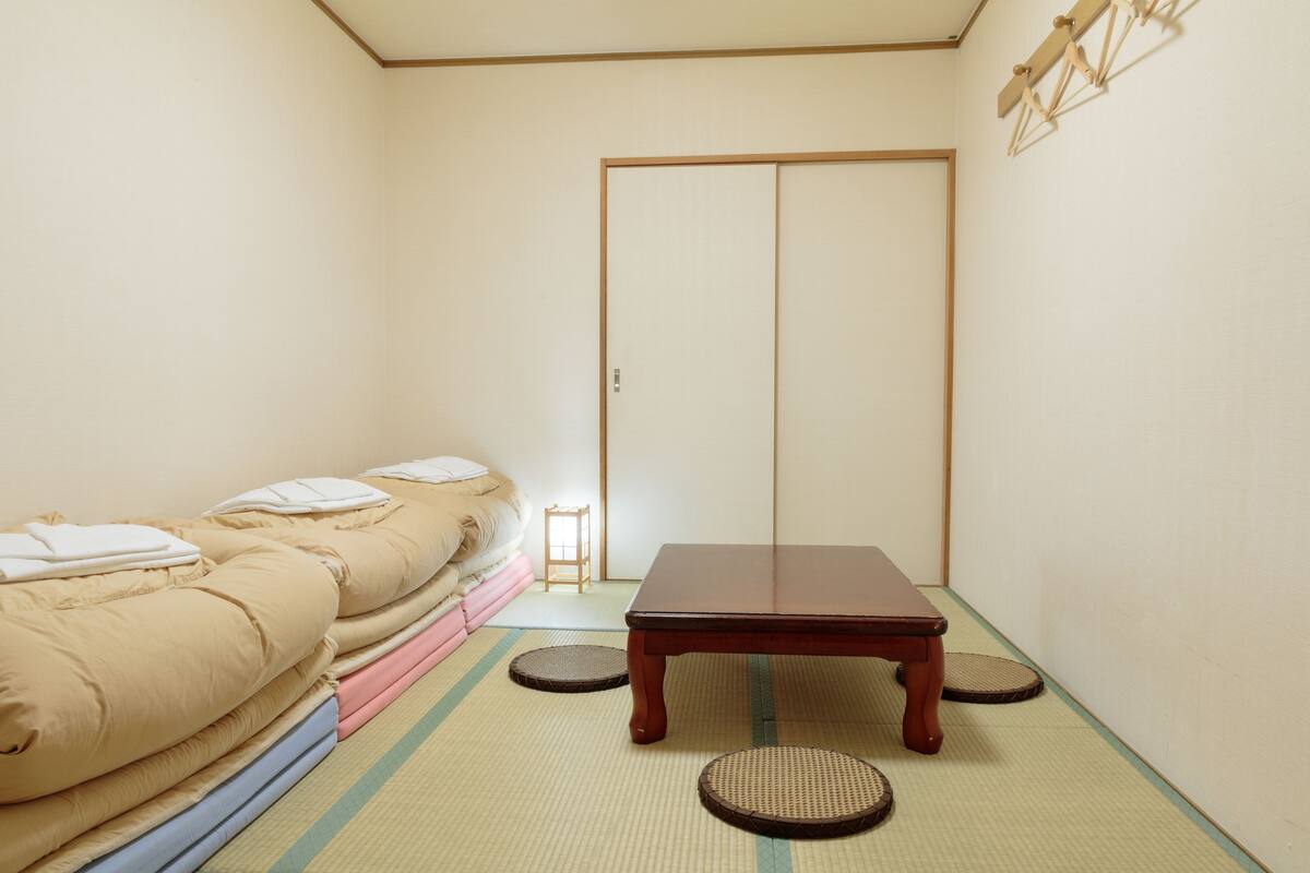 Ein traditioneller japanischer Raum mit Tatami-Bodenbelag, einem niedrigen Holztisch und einem Futonbett mit Kissen.