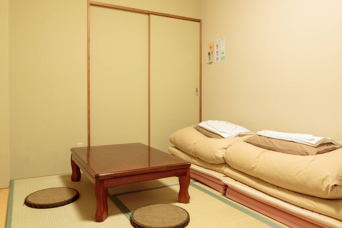 Ein Zimmer mit Tatami-Boden, einem niedrigen Holztisch, einer Couch mit Kissen und einer Schiebetür.