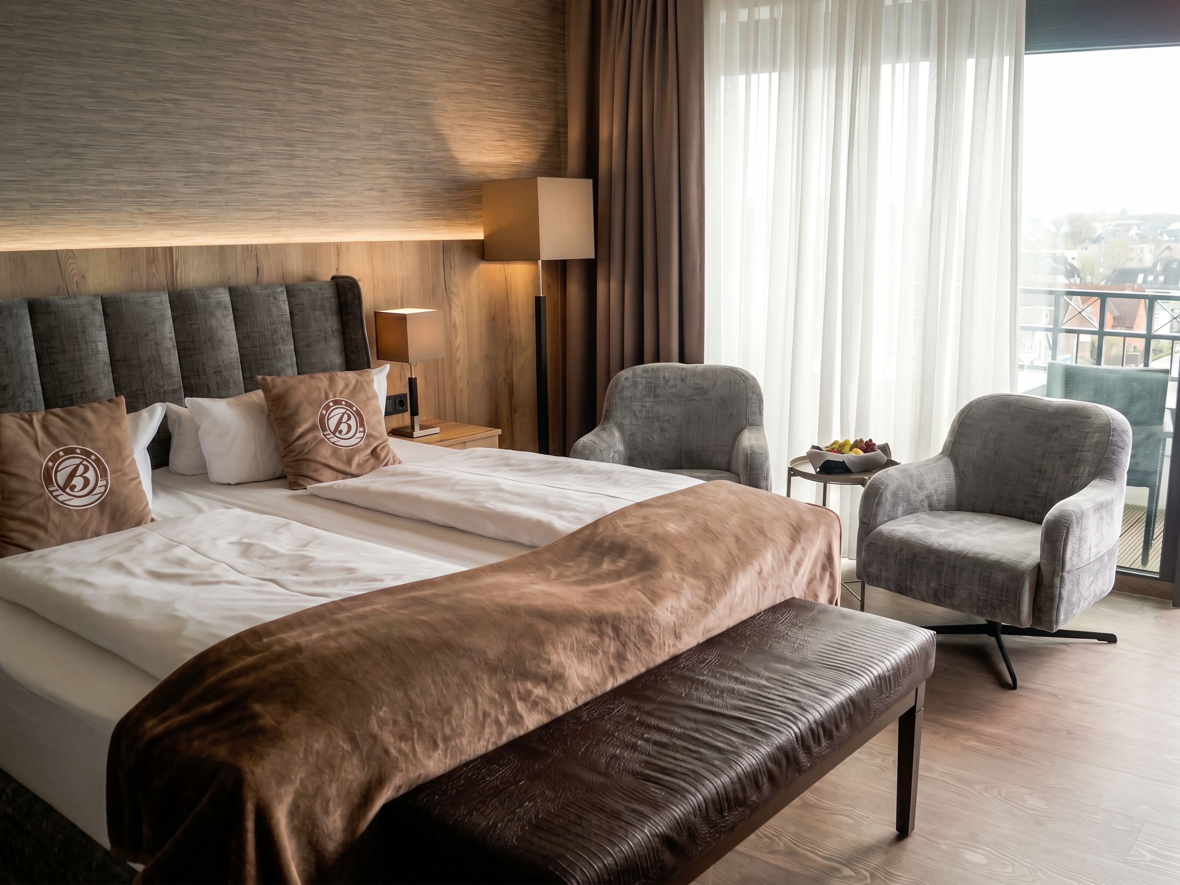 Ein Hotelzimmer mit einem großen Bett, einer Bank, zwei Sesseln, einem kleinen Tisch mit Obst und Blick auf die Stadt durch das Fenster.
