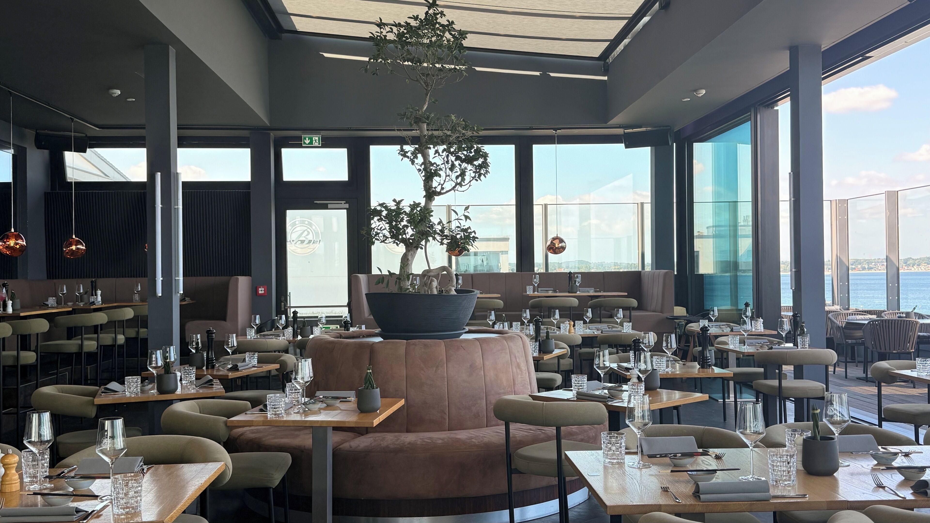 A modern restaurant with round tables, cushioned seating, and large windows offering a view of the sea.