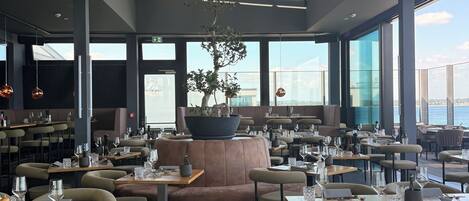 A modern restaurant with round tables, cushioned seating, and large windows offering a view of the sea.