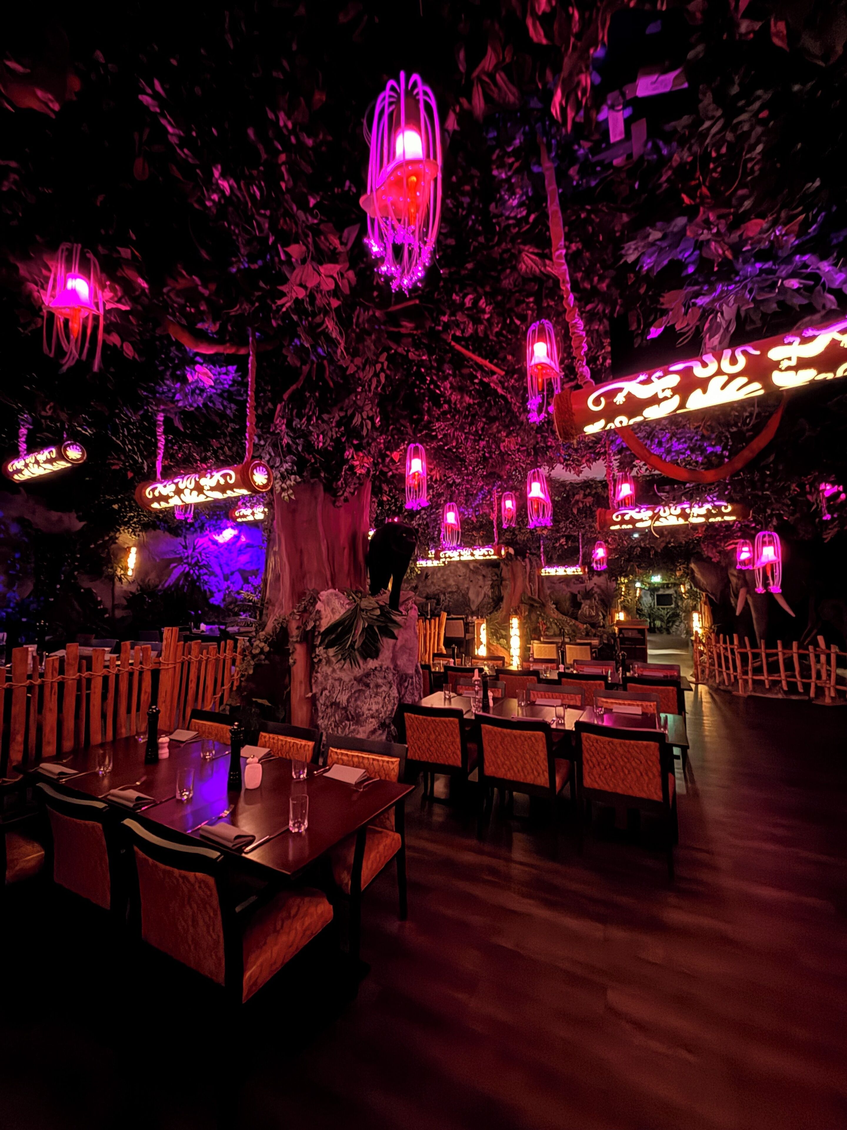 A dimly lit restaurant with wooden tables and chairs, hanging lanterns, and a backdrop of foliage.