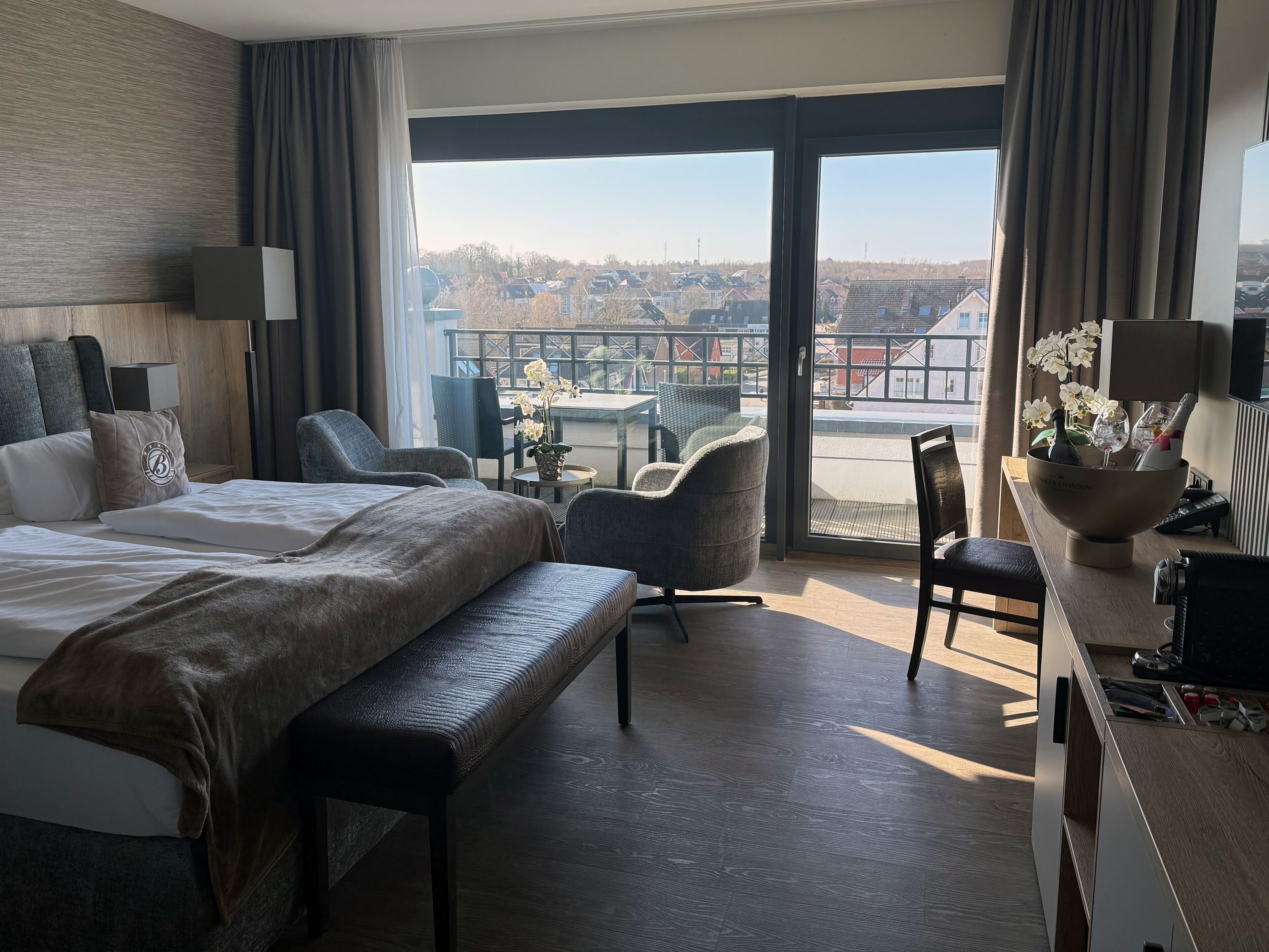 Ein Hotelzimmer mit einem großen Bett, einem Schreibtisch, einem Stuhl und einem Balkon mit Blick auf Gebäude.