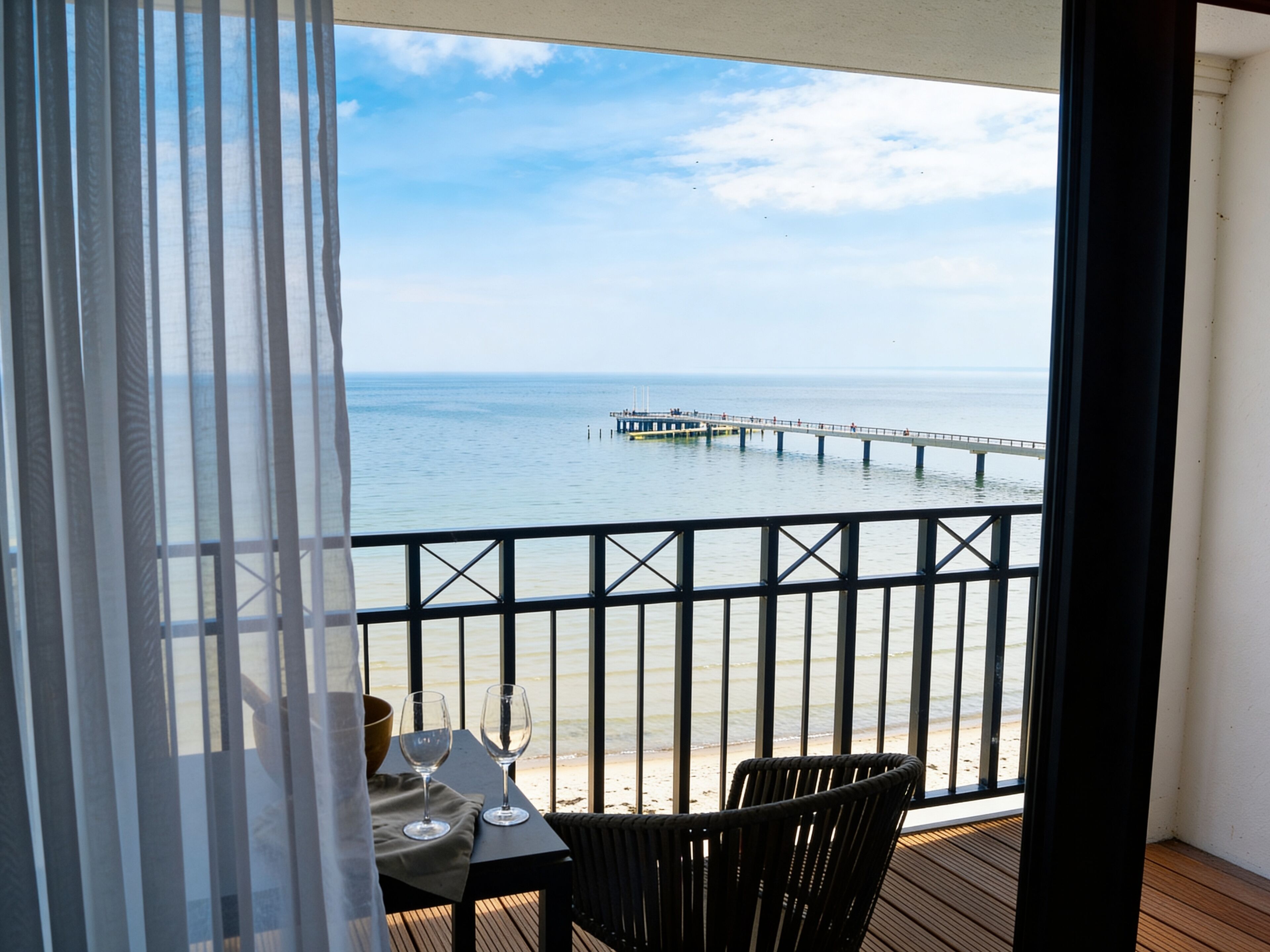 A balcony with a view of the sea and a pier. There is a table with two glasses and a bowl.