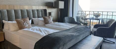 Habitación de hotel con una cama grande, un banco, una silla y vista al mar desde un balcón.