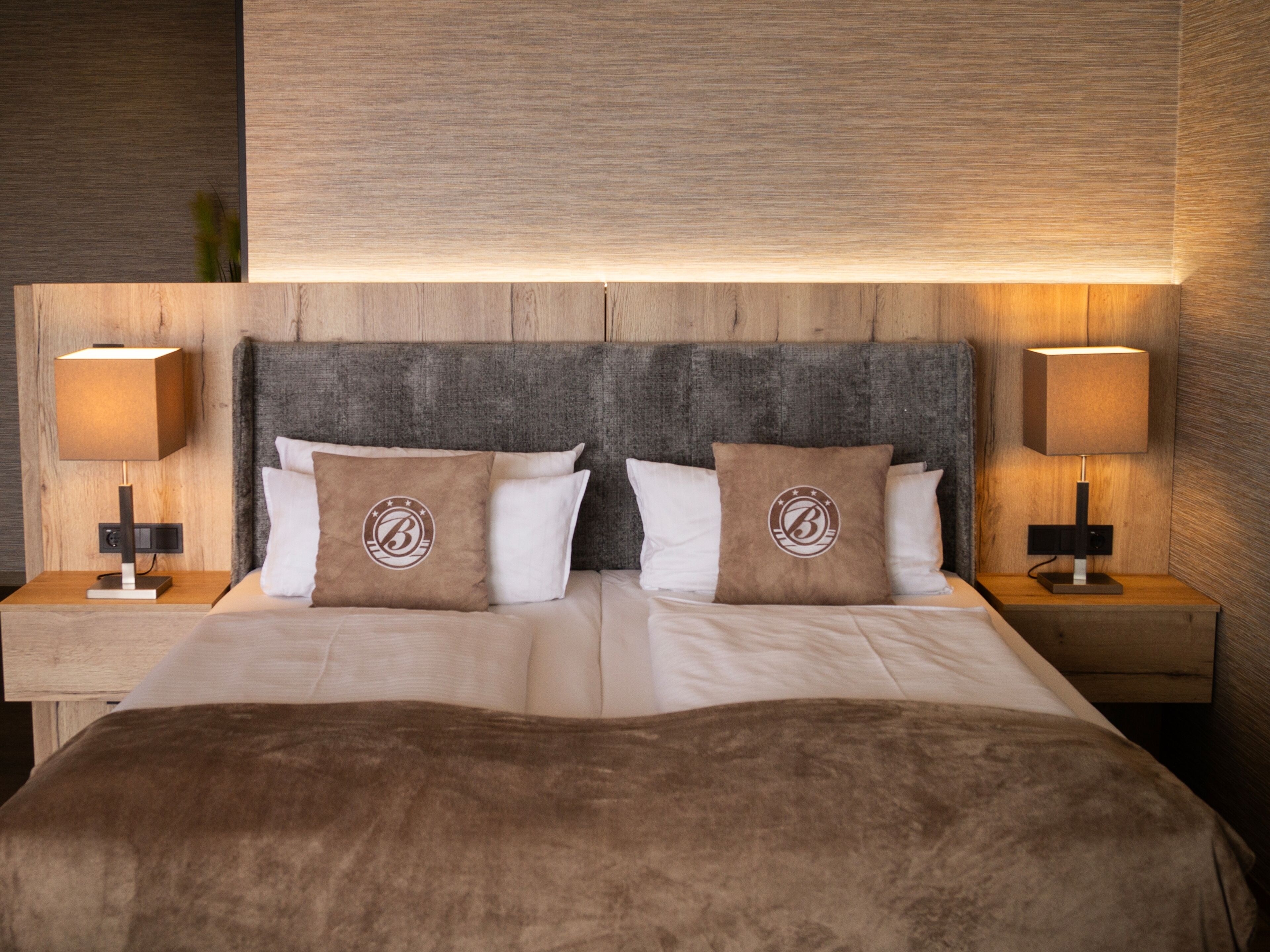 Cama doble con cabecera de madera, dos lámparas de noche y dos almohadas decorativas con un logotipo.