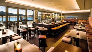 A modern bar area with a long counter, stools, and tables, overlooking a body of water. - Bayside (Scharbeutz)