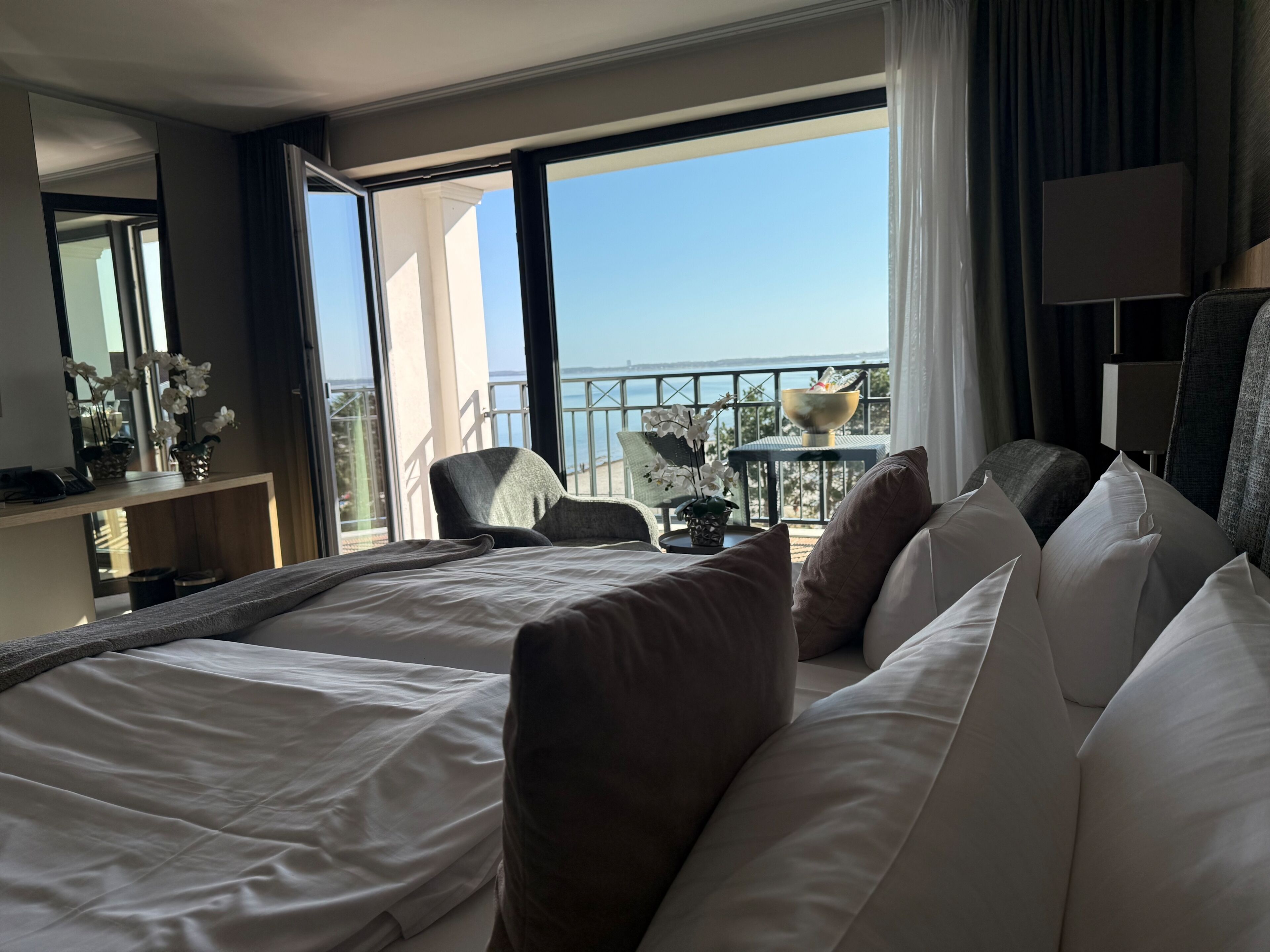 A hotel room with a bed, a chair, a small table with a bowl, and a balcony with a view of the sea.
