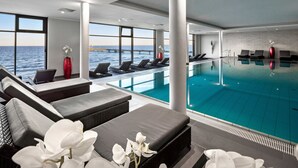 A modern indoor swimming pool with lounge chairs and sofas, large windows overlooking the sea, and a view of the coastline. - Bayside (Scharbeutz)