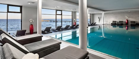 En moderne indendørs swimmingpool med liggestole og sofaer, store vinduer med havudsigt og kystlinjen.
