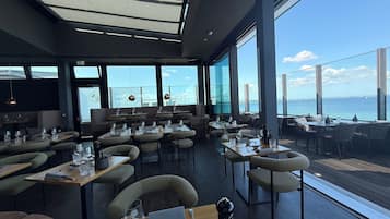 A restaurant with tables and chairs arranged for dining, large windows offering a view of the sea, and a modern interior design.