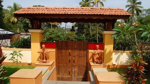 A wooden gate with two statues on either side, a tiled roof structure above, and a well-maintained garden with palm trees and potted plants. - Martin's Comfort (Betalbatim)
