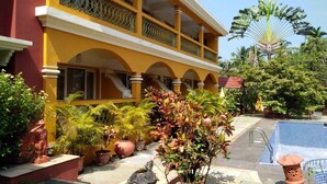A two-story building with a yellow facade, a balcony with ornate railings, and a pool area with a blue tiled deck. - Martin's Comfort (Betalbatim)