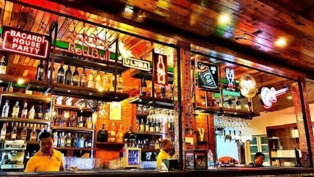 A bar with wooden shelves stocked with bottles, a neon sign for 'BACARDI HOUSE PARTY', and a 'REALBULL' sign. - Martin's Comfort (Betalbatim)