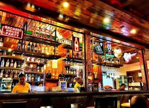 A bar with wooden shelves stocked with bottles, a neon sign for 'BACARDI HOUSE PARTY', and a 'REALBULL' sign. - Martin's Comfort (Betalbatim)