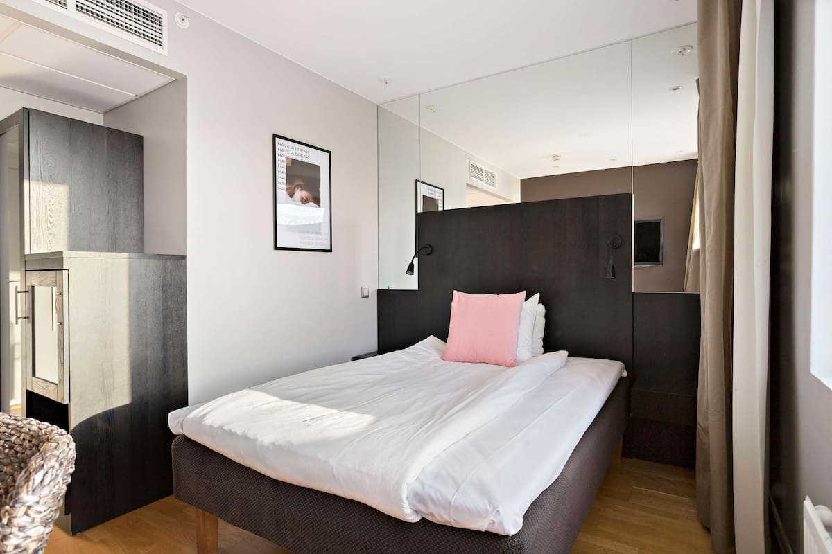 Moderni hotellihuone, jossa on suuri sänky, vaatekaappi, peili ja pieni ikkuna.