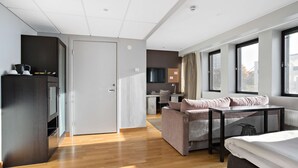 Ein modernes Wohnzimmer mit einer Couch, einem kleinen Tisch und einem Bett. Im Hintergrund ist eine Küchenzeile mit einem Fernseher und einem Fenster zu sehen.