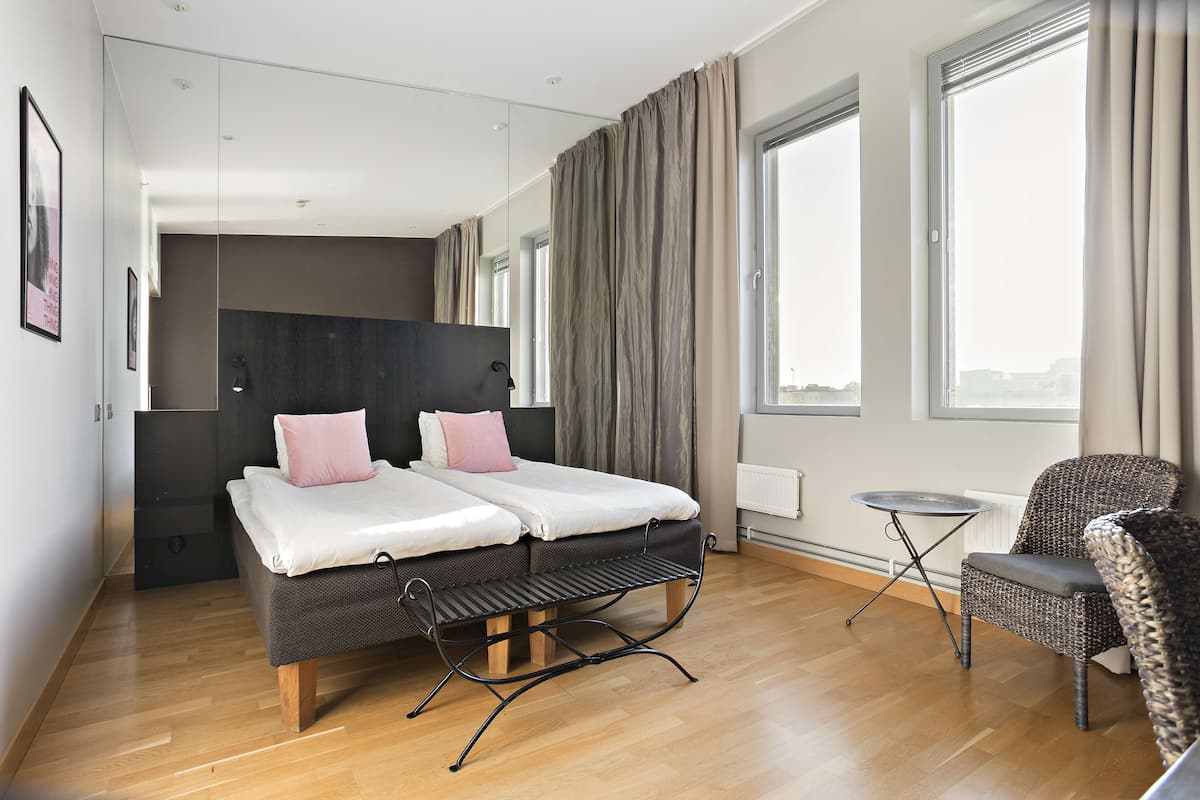 Ein modernes Hotelzimmer mit einem Doppelbett, einem kleinen Nachttisch, einem Korbstuhl und großen Fenstern.