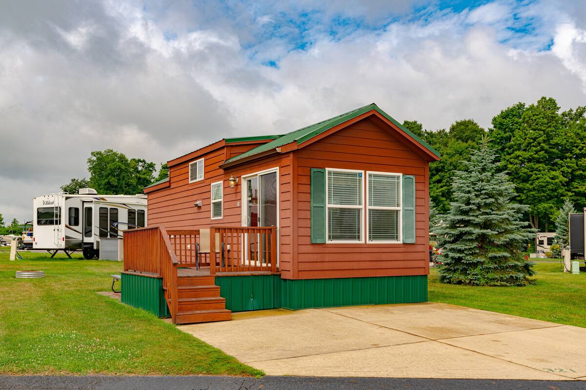 A small, single-story cabin with a green porch and a red roof, surrounded by grass and trees.