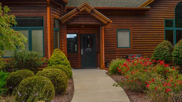 A log cabin with a covered porch, a chimney, and a well-maintained garden with various plants and flowers. - Sun Retreats Gun Lake (Hopkins)