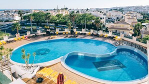 Una amplia piscina curva con tumbonas y sombrillas, rodeada de una zona ajardinada junto a la piscina con vistas a la ciudad al fondo.