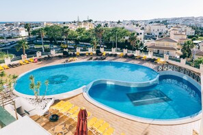A large, curved swimming pool with sun loungers and umbrellas, surrounded by a poolside area with a view of the city in the background.