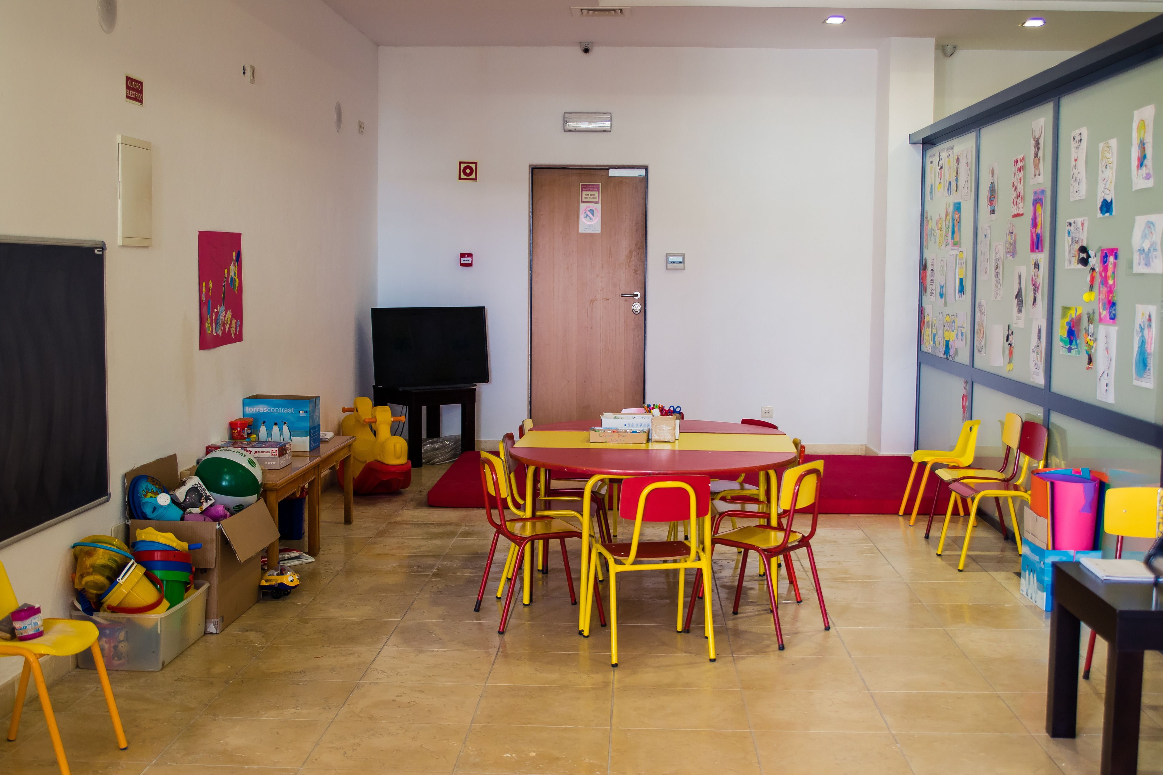 A classroom with a red table, yellow chairs, a blackboard, and children's artwork on the wall.