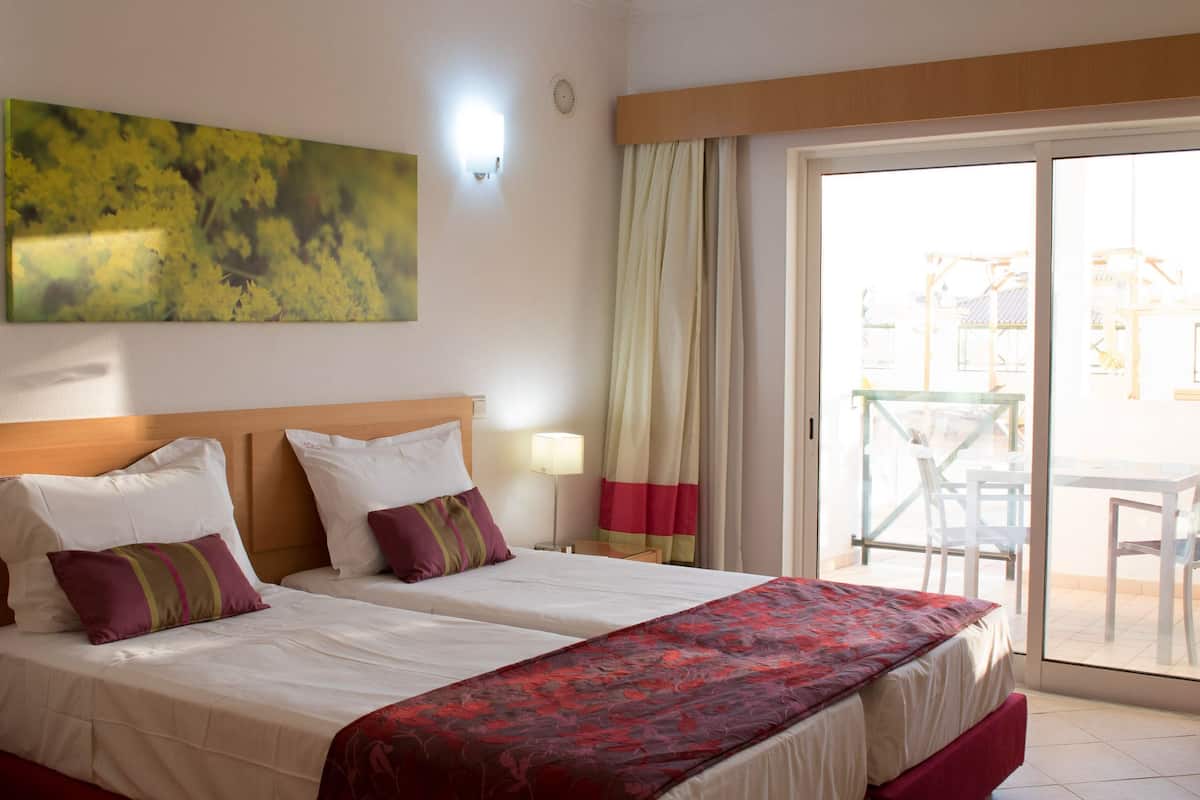 Une chambre d’hôtel avec deux lits, une grande fenêtre, un balcon avec une table et des chaises, et un tableau au mur.